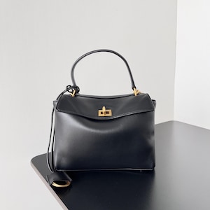 May include: A black leather handbag with a top handle and a rectangular shape. The bag features a gold-colored clasp and hardware. A small, rectangular tag hangs from the side. The bag is sitting on a black surface.
