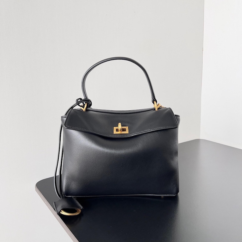 May include: A black leather handbag with a top handle and a rectangular shape. The bag features a gold-colored clasp and hardware. A small, rectangular tag hangs from the side. The bag is sitting on a black surface.