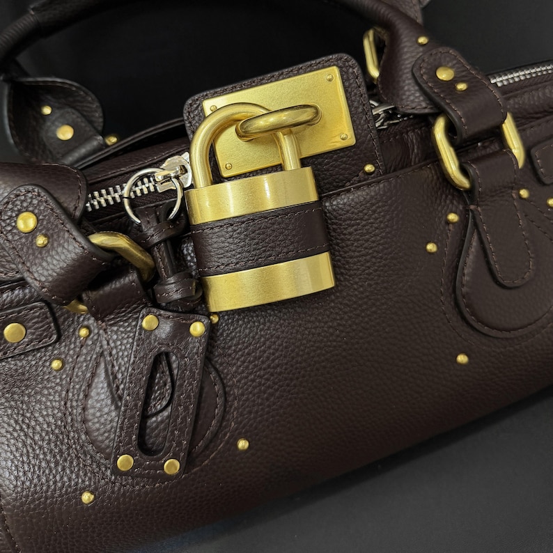 May include: A dark brown leather handbag with gold-toned hardware, including a padlock closure. The bag features a textured surface and is adorned with small, gold studs. The zipper and other metal accents are also gold.