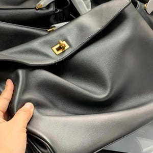 May include: Black leather handbag with a gold-colored clasp. The bag has a flap closure and a smooth texture. Several other black bags are visible in the background.