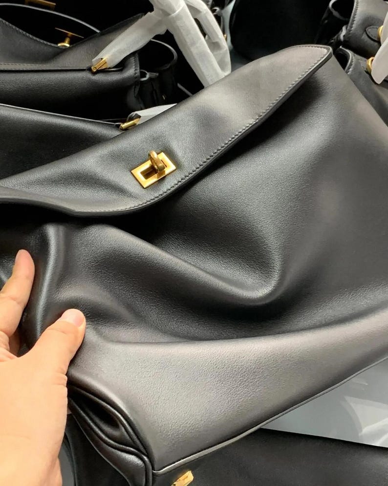 May include: Black leather handbag with a gold-colored clasp. The bag has a flap closure and a smooth texture. Several other black bags are visible in the background.
