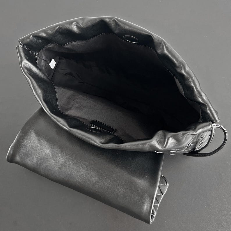 May include: Black leather handbag with an open top, revealing a spacious interior. The bag features a woven design on the side and a flap closure. The bag is shown against a dark background.
