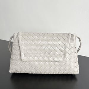 May include: A light gray, woven leather clutch bag with a flap closure. The bag features a rectangular shape with a woven pattern and thin shoulder straps. The bag is approximately 12 inches wide.
