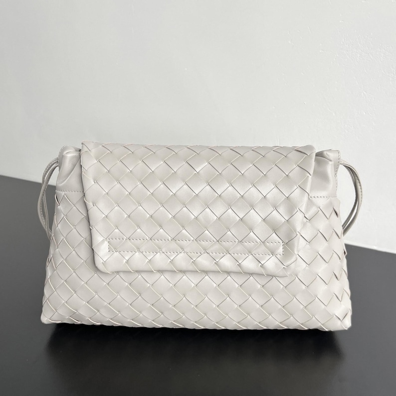 May include: A light gray, woven leather clutch bag with a flap closure. The bag features a rectangular shape with a woven pattern and thin shoulder straps. The bag is approximately 12 inches wide.