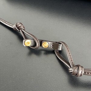 May include: Close-up of a dark brown leather strap with gold-colored metal rivets. The strap is knotted at both ends, and the rivets are visible on a small, rectangular piece of leather.