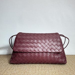 May include: A burgundy-colored woven leather clutch bag with a flap closure. The bag features a rectangular shape with a woven pattern and thin, dark burgundy straps. The bag is set against a neutral background.