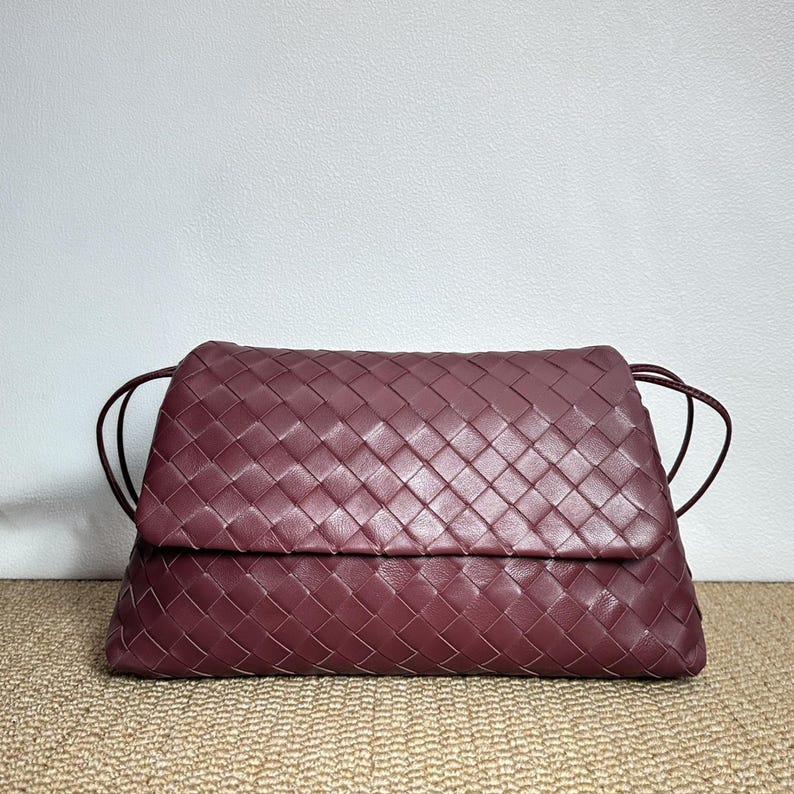 May include: A burgundy-colored woven leather clutch bag with a flap closure. The bag features a rectangular shape with a woven pattern and thin, dark burgundy straps. The bag is set against a neutral background.