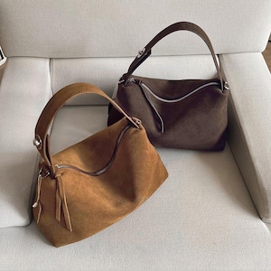 Könnte beinhalten: Zwei Wildlederhandtaschen in Brauntönen. Eine ist hellbraun, die andere ist dunkel schokoladenbraun. Beide haben einen geschwungenen Reißverschluss und einen einzelnen Griff. Die Taschen liegen auf einem hellen Sofa.