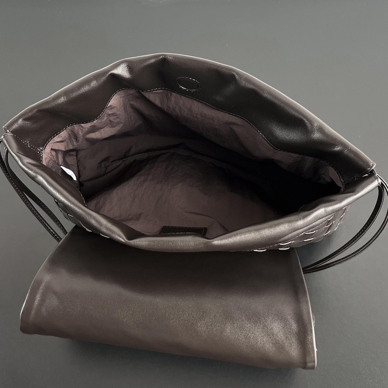 May include: Open view of a dark brown leather handbag. The bag features a woven design on the top flap and a drawstring closure. The interior is lined with a dark fabric. The bag is open, revealing the inside.