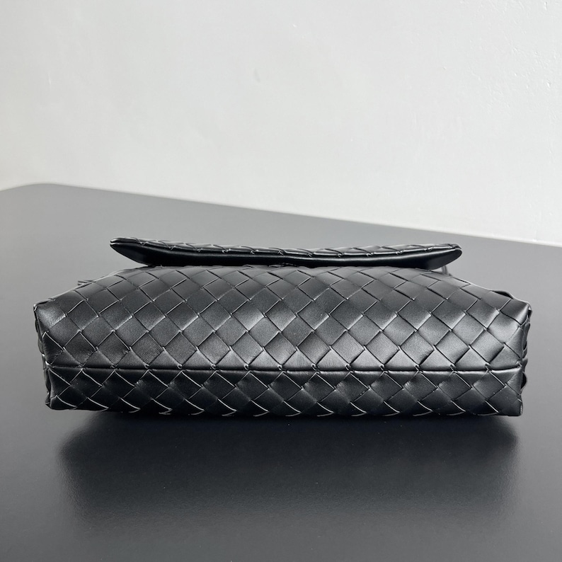 May include: A black leather clutch with a woven pattern. The rectangular bag has a flap closure and a textured surface. The bag is sitting on a black surface.