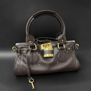 May include: A brown leather handbag with a top handle and gold-tone hardware. The bag features a lock closure with a key and small gold studs. The bag has a textured surface and a zipper closure.