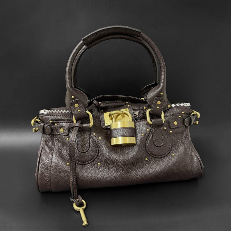 May include: A brown leather handbag with a top handle and gold-tone hardware. The bag features a lock closure with a key and small gold studs. The bag has a textured surface and a zipper closure.