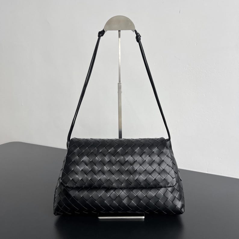 May include: Black woven leather shoulder bag with a flap closure. The bag features a rectangular shape with a woven pattern and a long, thin black strap. The bag is displayed on a silver stand against a white background.