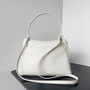 May include: A white leather handbag with a top handle and a detachable shoulder strap. The bag has a rectangular shape with a structured design and silver-tone hardware. The bag is sitting on a black surface.