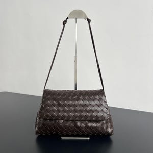 May include: A dark brown woven leather shoulder bag with a flap closure. The bag has a long, thin strap and is displayed on a silver metal stand against a white background.