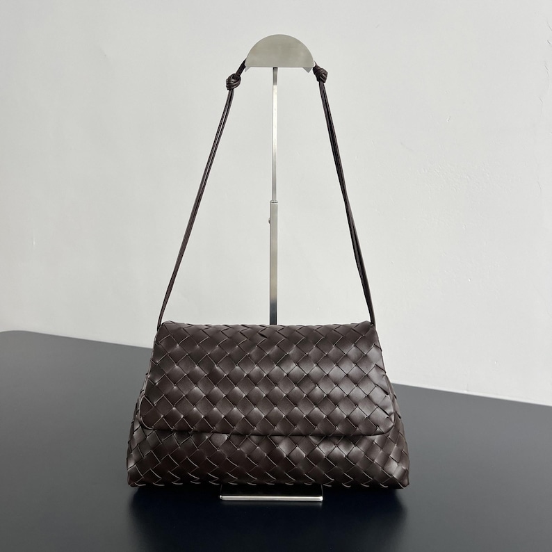 May include: A dark brown woven leather shoulder bag with a flap closure. The bag has a long, thin strap and is displayed on a silver metal stand against a white background.