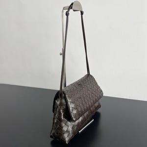 May include: A dark brown, woven leather crossbody bag with a long, thin strap. The bag features a flap closure and a rectangular shape. The leather has a textured appearance, and the bag is displayed on a black surface.