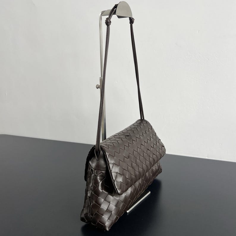 May include: A dark brown, woven leather crossbody bag with a long, thin strap. The bag features a flap closure and a rectangular shape. The leather has a textured appearance, and the bag is displayed on a black surface.