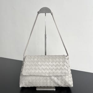 May include: A light gray, woven leather shoulder bag with a flap closure. The bag has a rectangular shape and a long, thin strap. The bag is displayed on a silver metal stand against a white background.