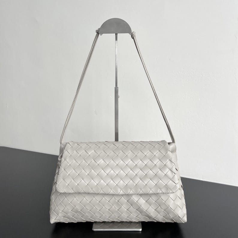May include: A light gray, woven leather shoulder bag with a flap closure. The bag has a rectangular shape and a long, thin strap. The bag is displayed on a silver metal stand against a white background.