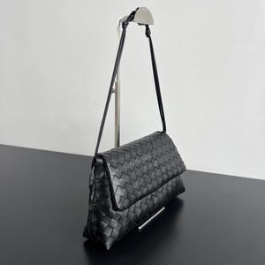 May include: Black woven leather shoulder bag with a flap closure. The bag has a long, thin strap and a rectangular shape. The bag is displayed on a silver metal stand against a white background.