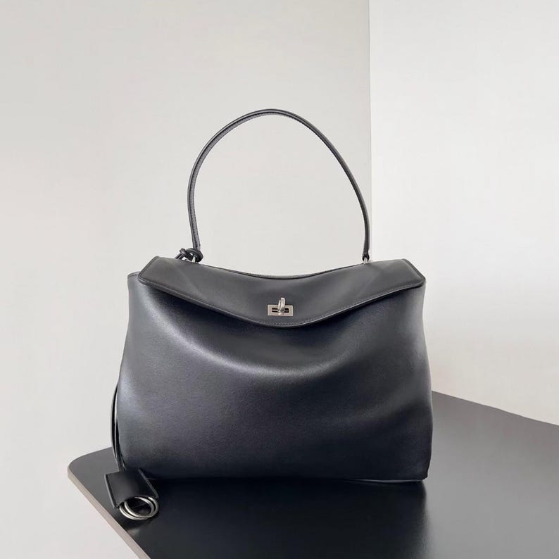 May include: Black leather handbag with a curved top handle and a silver-tone clasp. The bag has a structured shape with a flap closure and a small, dangling tag. The bag is sitting on a black surface.