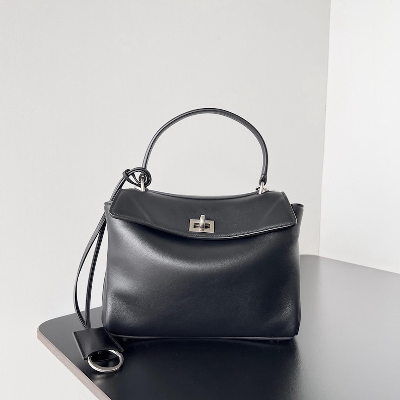 May include: Black leather handbag with a top handle and a detachable shoulder strap. The bag features a silver-tone clasp closure and a small, attached leather tag. The design is minimalist and elegant, suitable for various occasions.