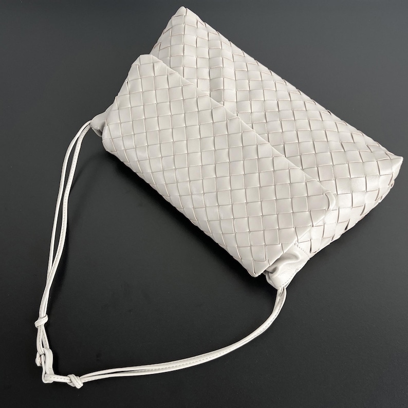 May include: A light gray woven leather clutch bag with a long, thin strap. The bag has a rectangular shape with a flap closure. The woven pattern is visible across the entire surface of the bag.