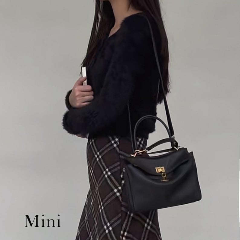 May include: Black leather handbag with a top handle and a shoulder strap. The bag has a gold-colored clasp and hardware. The word "Mini" is displayed at the bottom of the image. The bag is being carried.