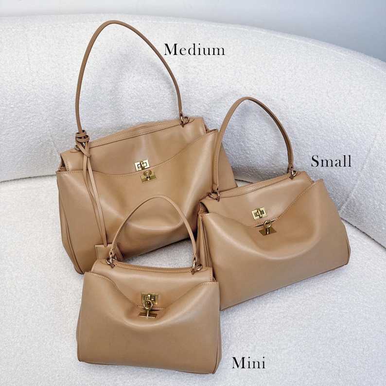 May include: Three tan leather handbags in varying sizes: medium, small, and mini. Each bag features a structured design with a top handle, a gold-tone clasp, and a rectangular shape. The bags are displayed on a white, textured surface.