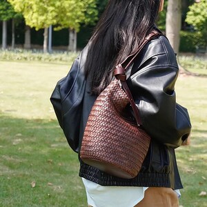 May include: A brown leather woven bucket bag with a long strap. The bag is worn over the shoulder of a person wearing a black leather jacket.