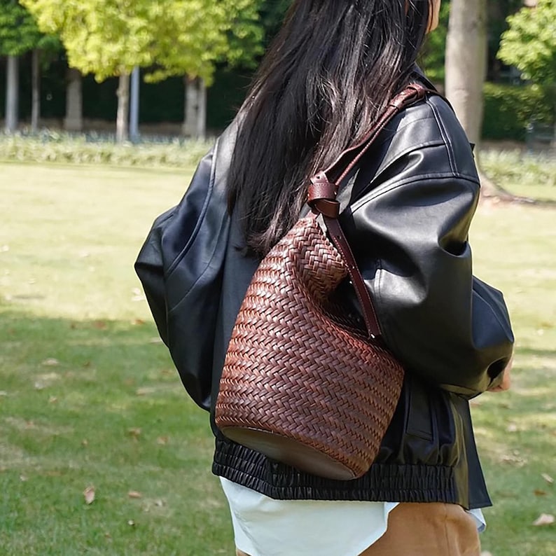 May include: A brown leather woven bucket bag with a long strap. The bag is worn over the shoulder of a person wearing a black leather jacket.