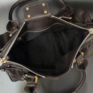 May include: Dark brown leather handbag with a textured finish. The bag features a top zipper closure, gold-toned hardware, and two top handles. The interior is lined with black fabric.