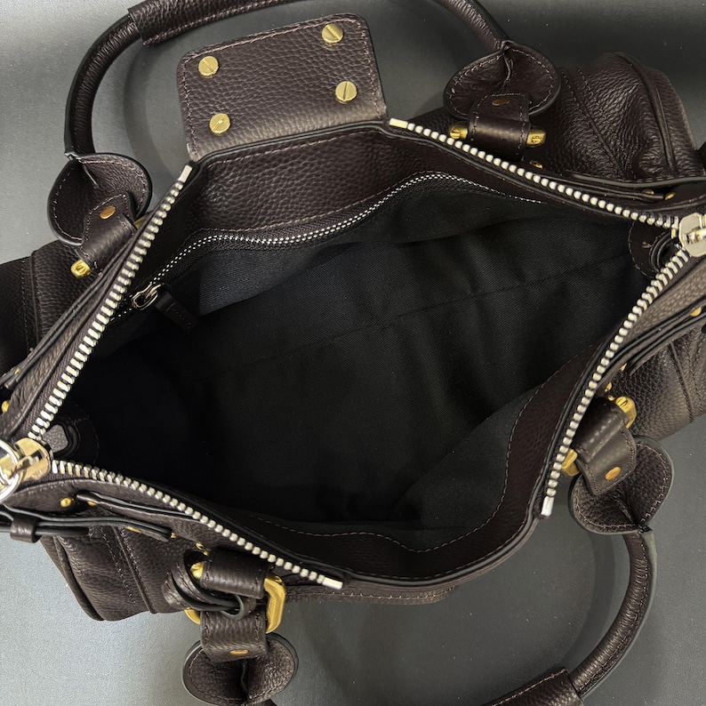 May include: Dark brown leather handbag with a textured finish. The bag features a top zipper closure, gold-toned hardware, and two top handles. The interior is lined with black fabric.