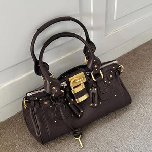May include: A dark brown handbag with a gold-colored lock and hardware. The bag has two handles and a zipper closure. Small gold studs are evenly spaced across the exterior. A key hangs from the lock.