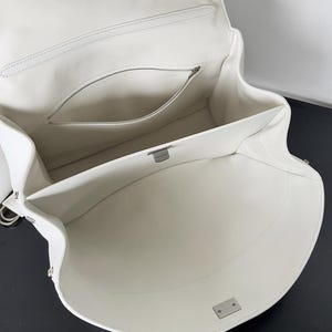May include: Open white leather handbag with a structured design. The interior features a zippered pocket and a spacious main compartment. Silver-tone hardware accents the bag, including a clasp closure and metal rings for a shoulder strap.
