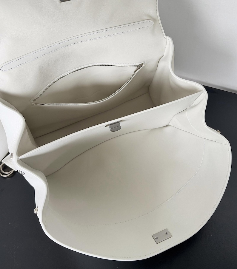 May include: Open white leather handbag with a structured design. The interior features a zippered pocket and a spacious main compartment. Silver-tone hardware accents the bag, including a clasp closure and metal rings for a shoulder strap.