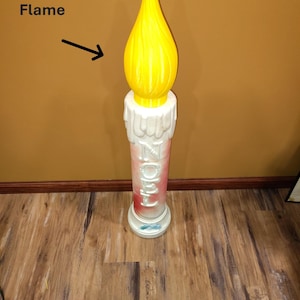 May include: A large decorative candle with a yellow flame and a white base. The word "NOEL" is printed vertically on the candle. The image also includes the text "Replacement Flame".