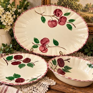 May include: A set of three cream-colored ceramic dishes featuring a fluted edge with a speckled maroon trim and hand-painted illustrations of dark red apples with green leaves on each piece, including one large oval platter and two bowls.