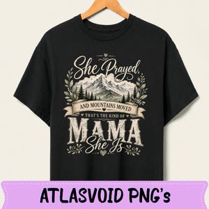 Puede incluir: Camiseta negra con el texto "She Prayed, And Mountains Moved" encima de "That's the kind of MAMA She Is". El diseño incluye una escena de montaña y detalles florales. La camiseta está colgada en una percha de madera.