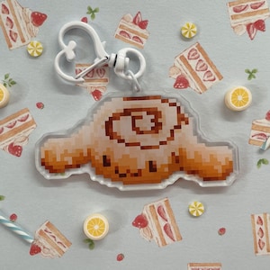 Puede incluir: Un llavero pixelado blanco y marrón con forma de perro de dibujos animados con un remolino en la cabeza. El llavero está sobre un fondo blanco con pequeños elementos decorativos como pasteles y limones.