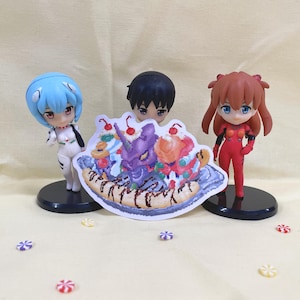 May include: A sticker featuring a banana split with various toppings, including whipped cream, cherries, and chocolate sauce. The sticker is surrounded by three anime figurines, one blue, one red, and one black.
