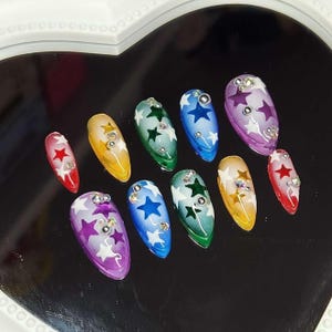 May include: A set of ten artificial nails with a star design. Each nail is a different color, including red, yellow, green, blue, and purple. The nails are decorated with white stars and rhinestones.