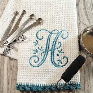 May include: White kitchen towel with a teal embroidered letter "A" and a teal crocheted border. The towel is folded and lying on a wooden surface.