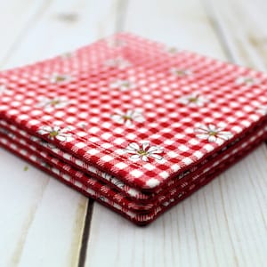 May include: A stack of four red and white gingham fabric coasters with a white daisy pattern. The coasters are folded and stacked on a white wooden surface.