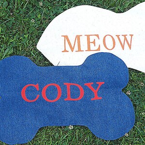 K&ouml;nnte beinhalten: Zwei Filzt&uuml;rmatten, eine blaue und knochenf&ouml;rmig mit dem Namen "Cody" in roten Buchstaben, die andere wei&szlig; mit dem Wort "MEOW" in orangefarbenen Buchstaben.