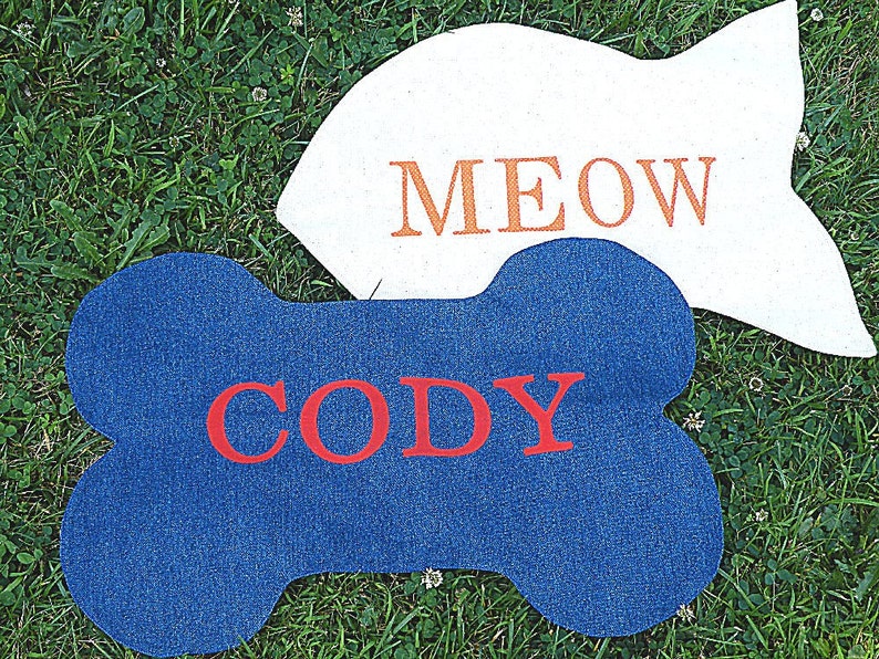 K&ouml;nnte beinhalten: Zwei Filzt&uuml;rmatten, eine blaue und knochenf&ouml;rmig mit dem Namen "Cody" in roten Buchstaben, die andere wei&szlig; mit dem Wort "MEOW" in orangefarbenen Buchstaben.
