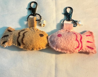 May include: Two felt fish keychains. One is tan with brown stripes, the other is pink with red details. Each keychain has a black metal clasp and a small white star charm. The fish are stitched together and appear to be handmade.