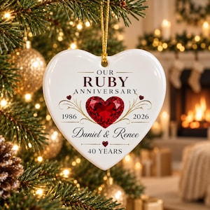 May include: A white heart-shaped ceramic ornament featuring a red gemstone graphic, the text "OUR RUBY ANNIVERSARY", "1986", "2026", "Daniel & Renee", and "40 YEARS". The ornament hangs from a gold string against a blurred Christmas tree background.