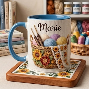 May include: A white ceramic mug with a blue handle and interior, featuring a basket of yarn and crochet hooks. The mug has the name "Marie" on it. It sits on a wooden coaster with a sunflower design. Books and craft supplies are in the background.